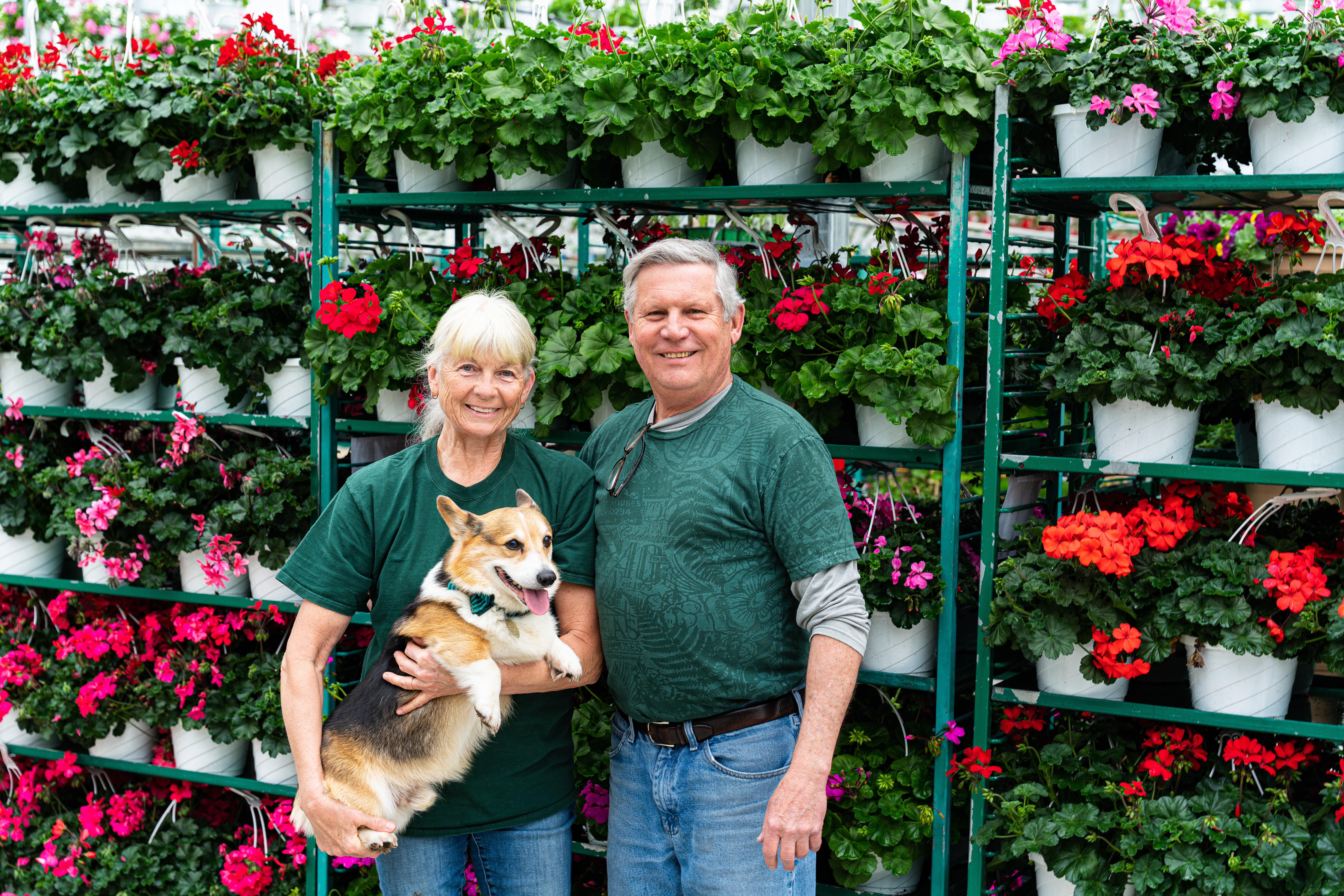 an image of the owners for the Lucas Greenhouses taken in front of potted flowers stacked on shelves with the lady holding a dog